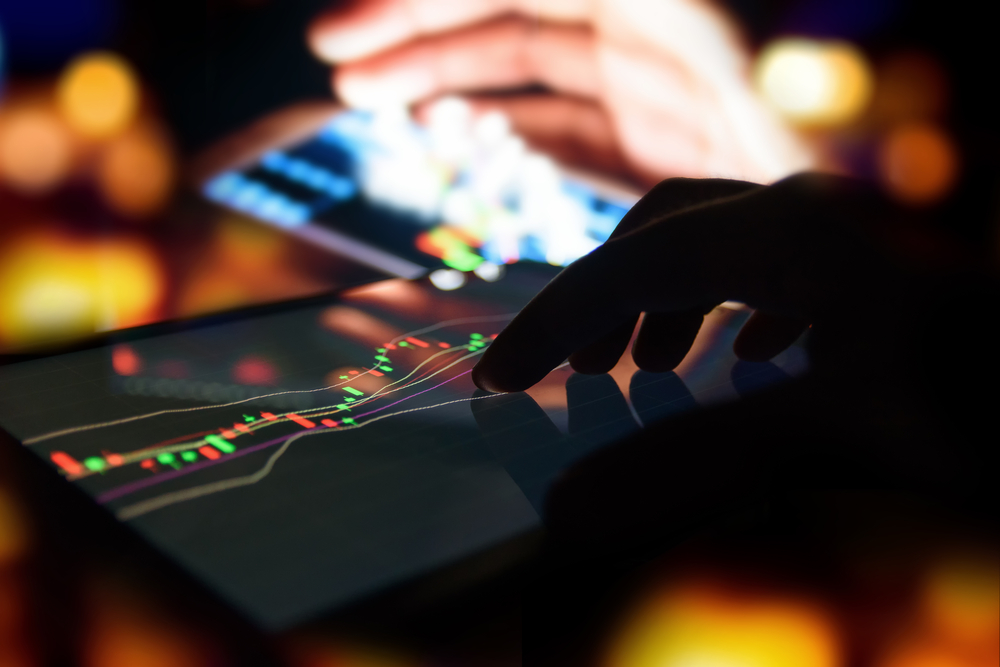 Hand taps a tablet showing a colorful stock candlestick chart in a dim, neon-lit setting.