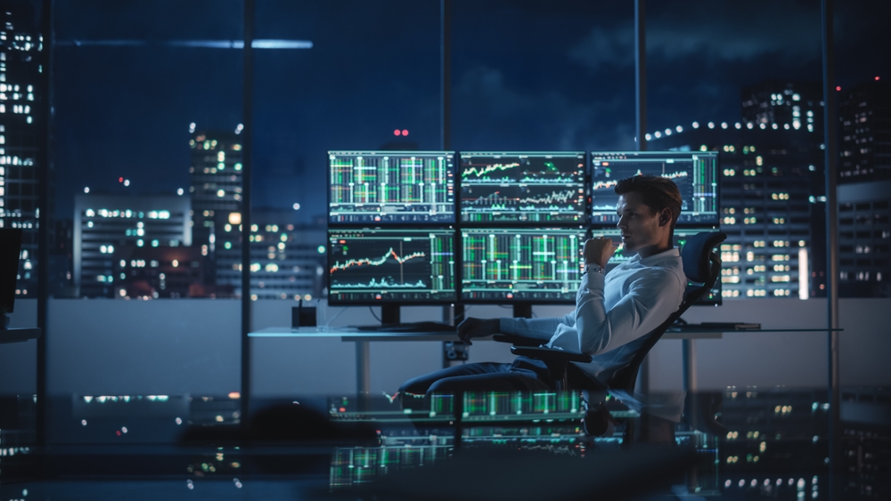 Man sits in a glass-walled office at night, monitoring multiple screens displaying stock charts and market data.